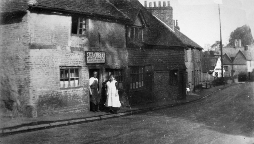 Benjamin Haylett stands in the sun outside his bootmaker's shop with his wife Isabell. It would have been very gloomy working the leather in the cottage with so few windows. Darenth Nursery shop can be seen further down the hill with its distinct white frontage and cat-slide roof. Tucked in behind the furthest of these cottages was a blacksmith's forge which was working until the 1920s.